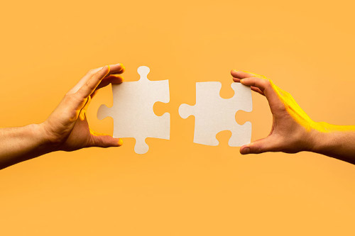 Zwei Hände halten leere Puzzlestücke vor einem orangefarbenen Hintergrund.